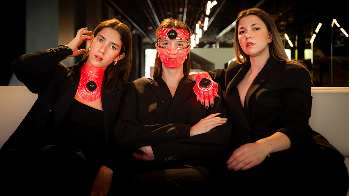 Three young woman in black wearing a red light mask for face, hands and neck respectively